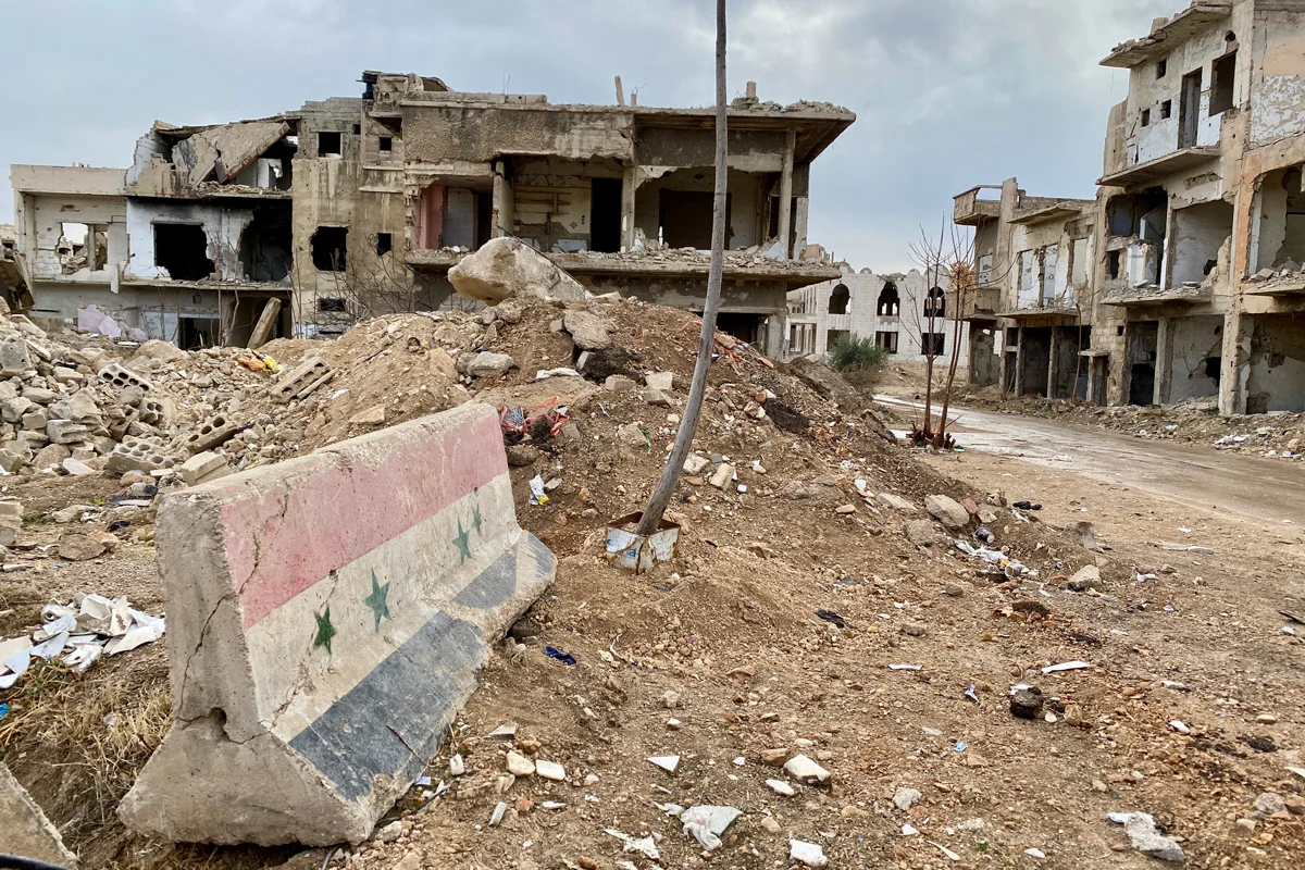 Остатки бетонного блока в цветах прежнего флага Сирии на фоне разрушенного здания, блоки использовались повсеместно на правительственных блокпостах в Сирии